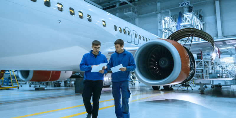 Commercial airliner: two technicians walk and compare inspection papers beside a large jet engine inside a brightly lit maintenance hangar with scaffolding.