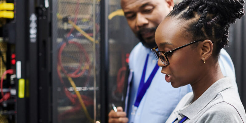 A female IT technician explains and points at server equipment while a male colleague observes; they stand beside a locked server rack filled with network cables in a data center.