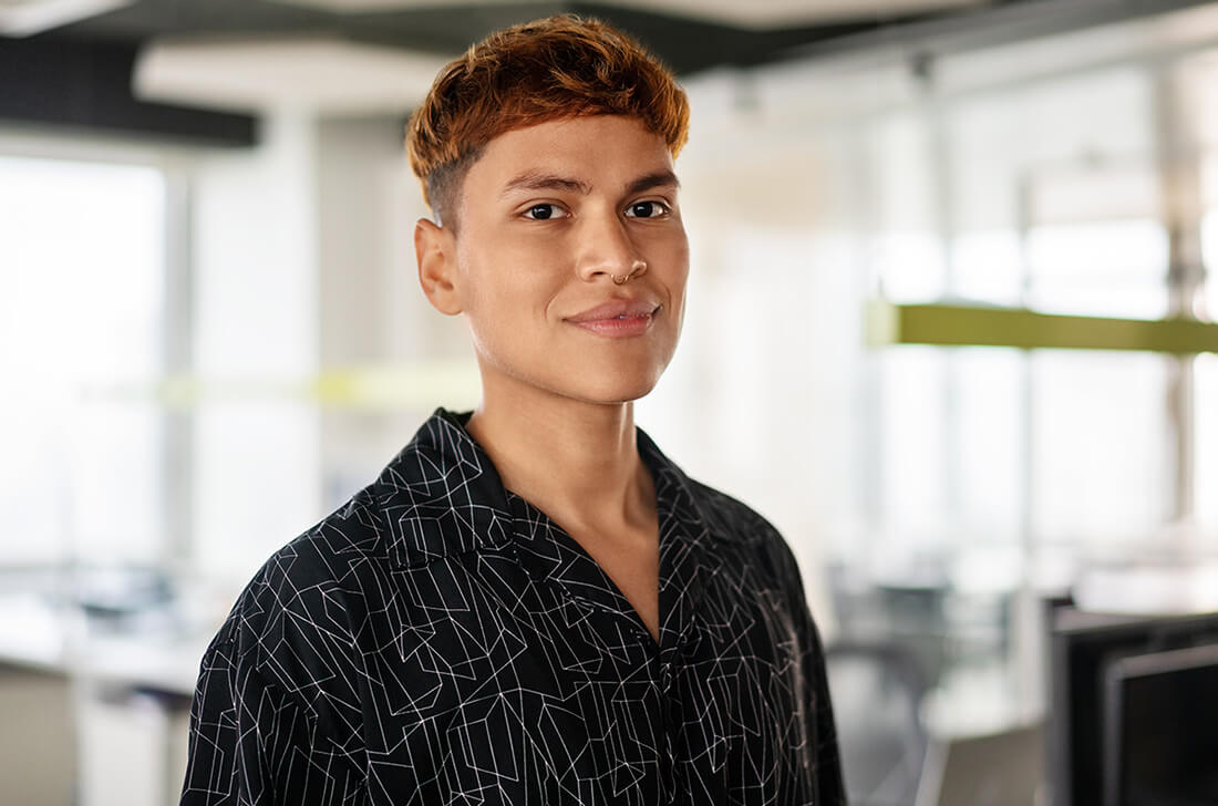 Person with short hair and geometric shirt smiles in modern office space.