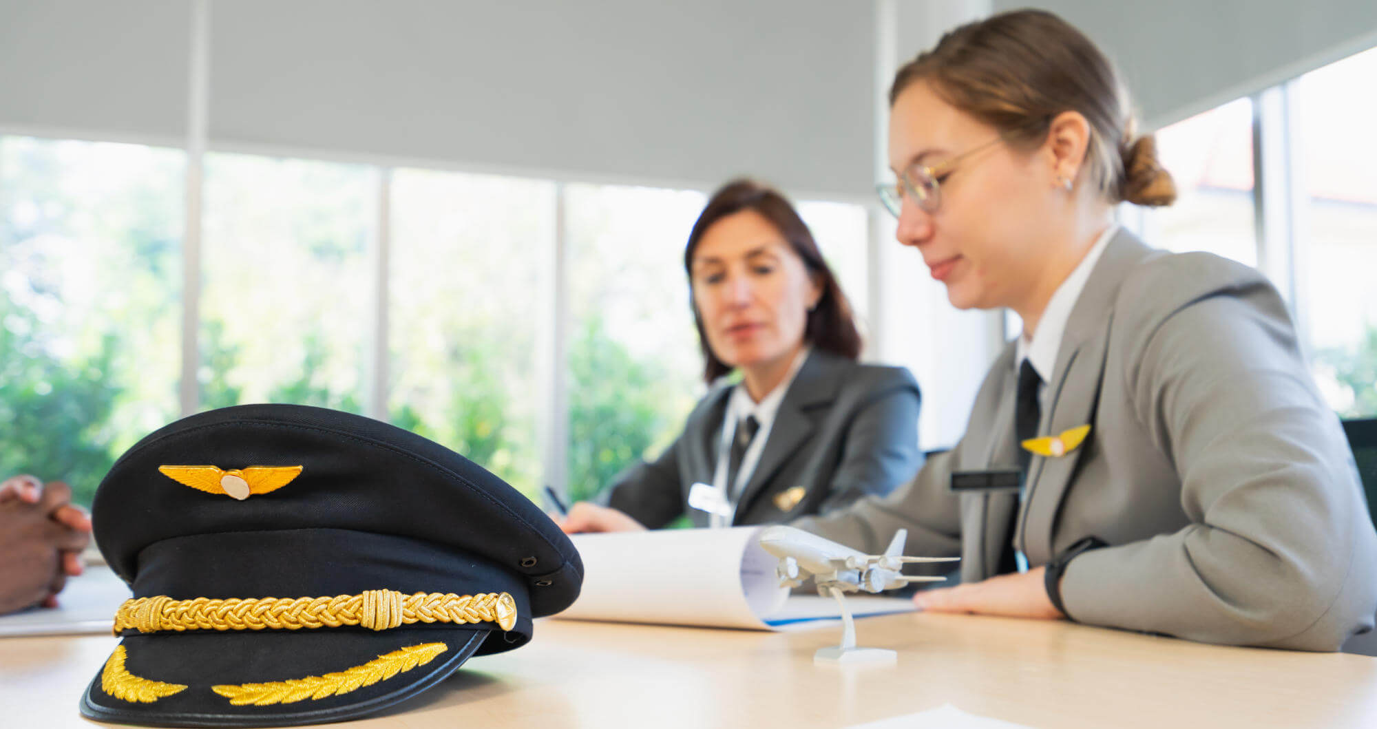 Pilot’s cap rests on a conference table while two uniformed crew members review documents beside a small model airplane in a bright office with large windows.