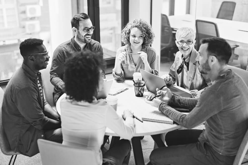 Six coworkers sit around a round table, talking and smiling, using laptops, notebooks and coffee in a bright, modern office meeting room.
