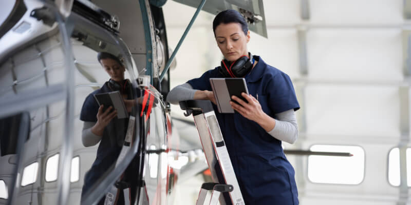 Technician examines a tablet while standing on a ladder beside a polished aircraft fuselage inside a spacious hangar; headset rests around her neck, the fuselage reflecting her image.