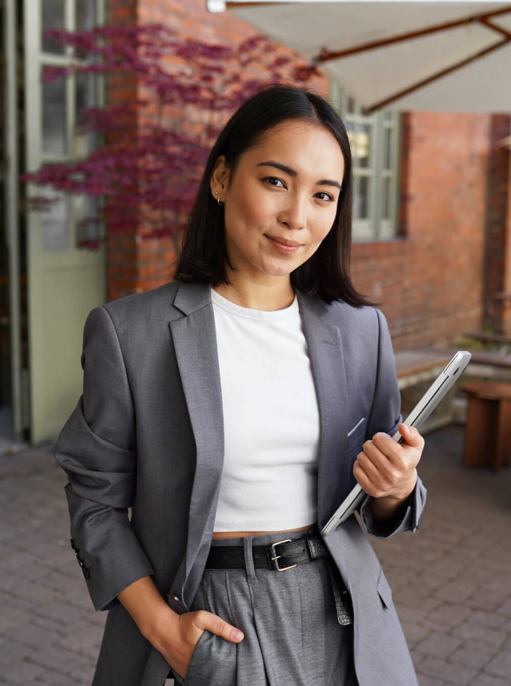 Woman in a gray suit holding a closed laptop under her arm, smiling gently while standing in a brick courtyard with a patio umbrella and a small ornamental tree.
