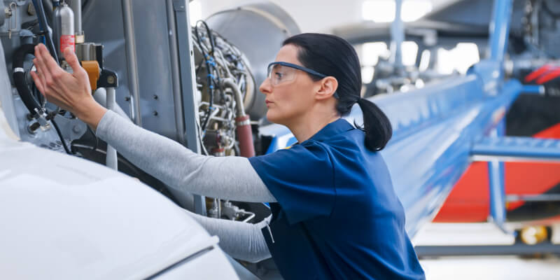 Aircraft engine being inspected by a technician adjusting components while wearing safety glasses and a navy work shirt, inside a spacious, well-lit maintenance hangar beside other aircraft parts.