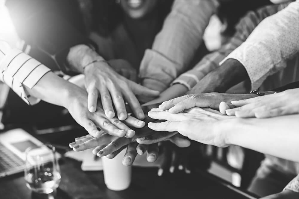 Multiple hands stacked in a team huddle over a reflective conference table with a coffee cup, glass, and laptop, suggesting collaboration in an office setting.
