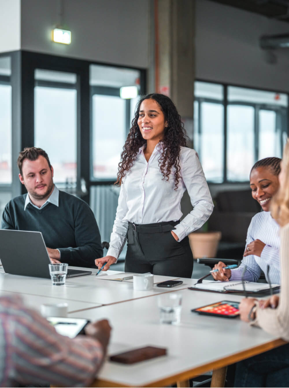 A woman in a white blouse stands leading a meeting, gesturing with a pen; colleagues sit around a table with laptops and tablets in a bright office with large windows.