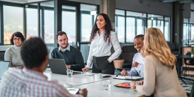 A woman in a white blouse stands leading a meeting, gesturing with a pen; colleagues sit around a table with laptops and tablets in a bright office with large windows.