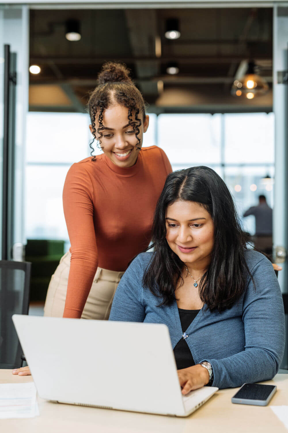 A seated woman working on a laptop while a colleague leans over, smiling and offering help, in a bright modern office with large windows and overhead pendant lights.