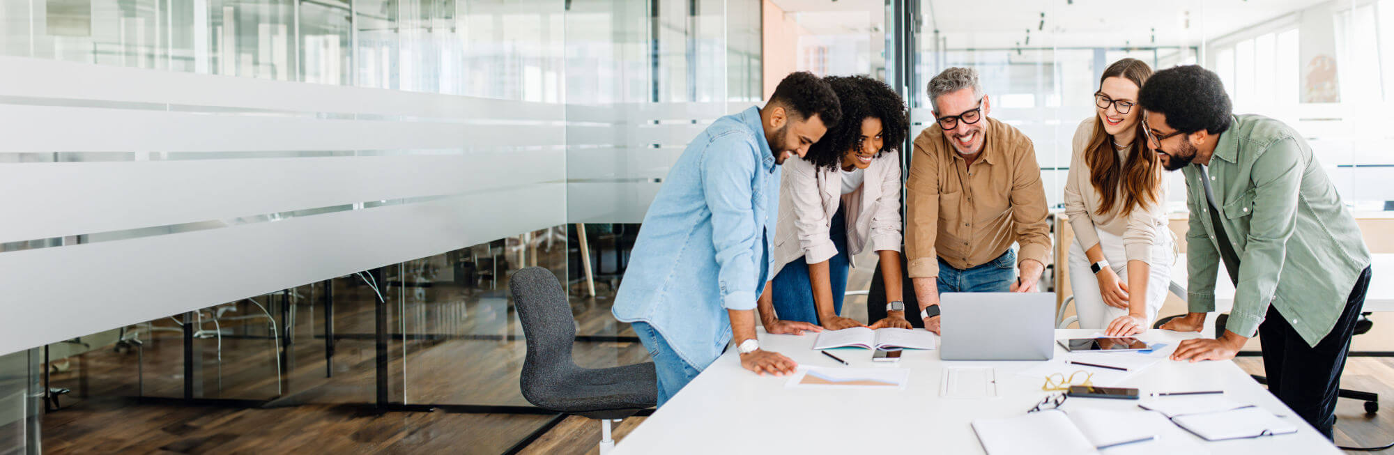 Five colleagues lean over a table, smiling and pointing at a laptop while discussing documents and tablets in a bright, glass-walled office meeting room.