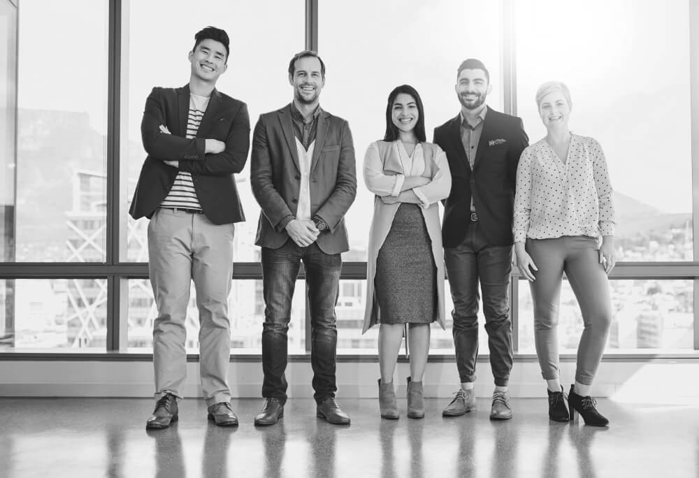 Five professionals stand smiling, three with arms crossed, in a sunlit high-rise office with floor-to-ceiling windows overlooking the city.