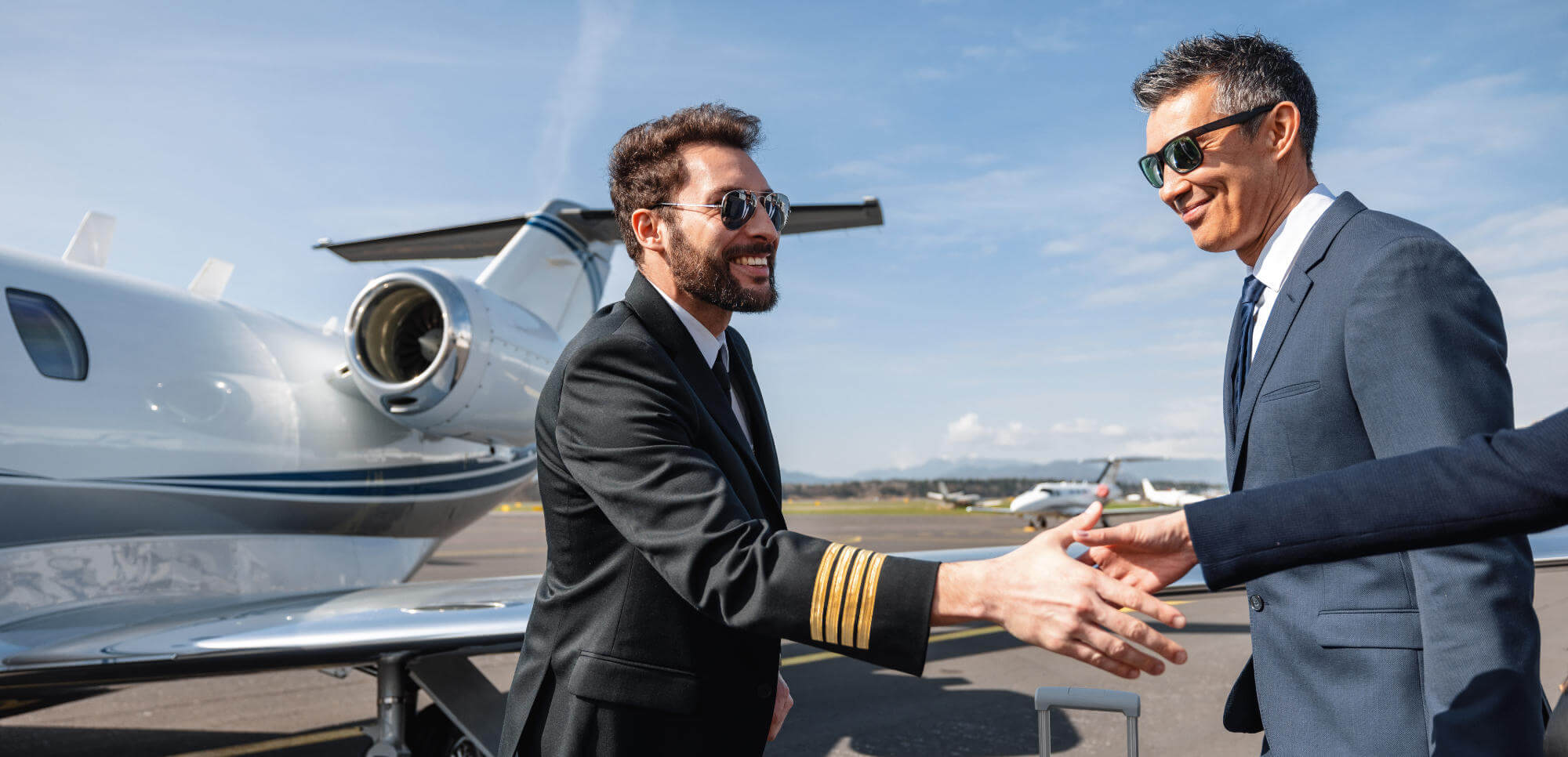 A pilot in a captain's uniform extends his hand to shake a suited businessman beside a parked private jet on an airport tarmac under a clear blue sky.