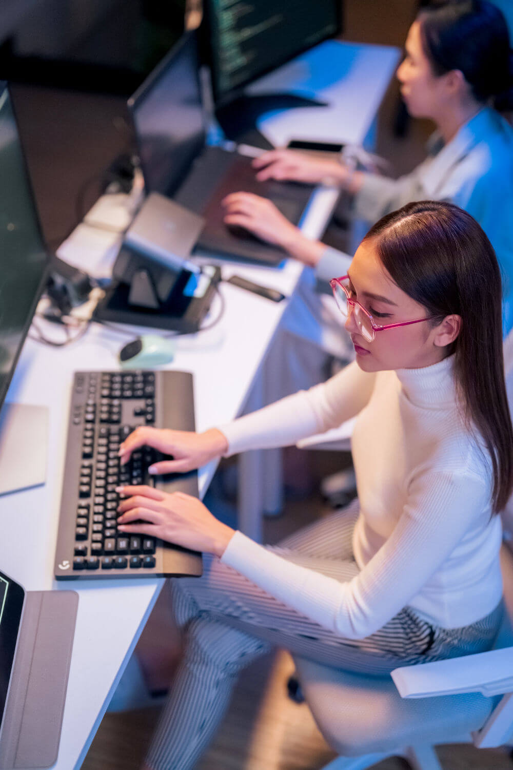 Young woman with pink glasses types on a desktop keyboard while seated at a white desk, multiple monitors and a colleague also working nearby in a dim, blue-lit office.
