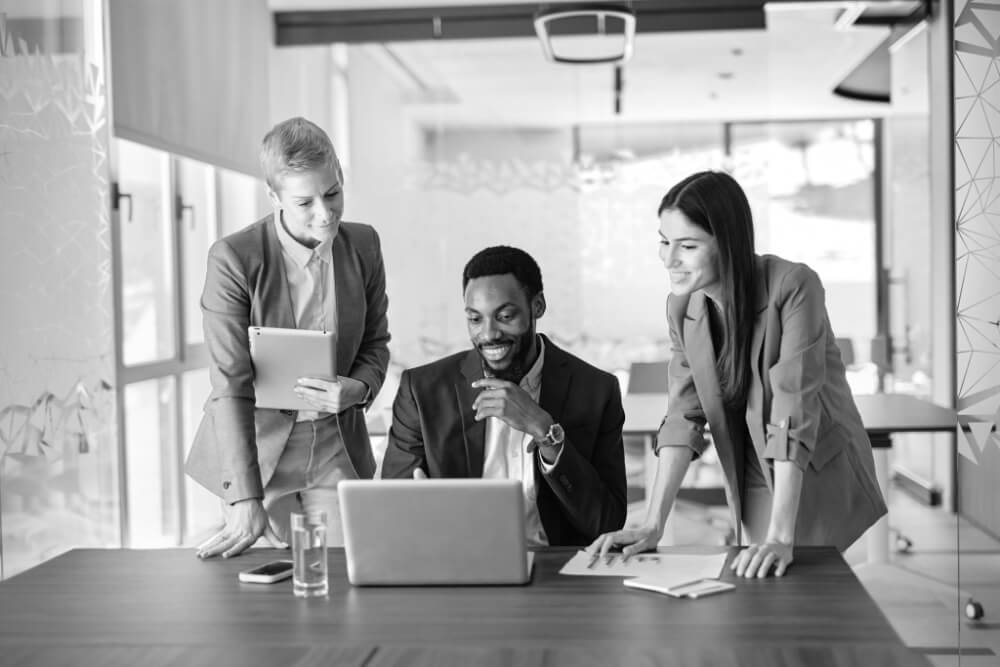 Three colleagues lean over a laptop, smiling and discussing a document; one holds a tablet, papers and a water bottle sit on the conference table in a glass-walled modern office.