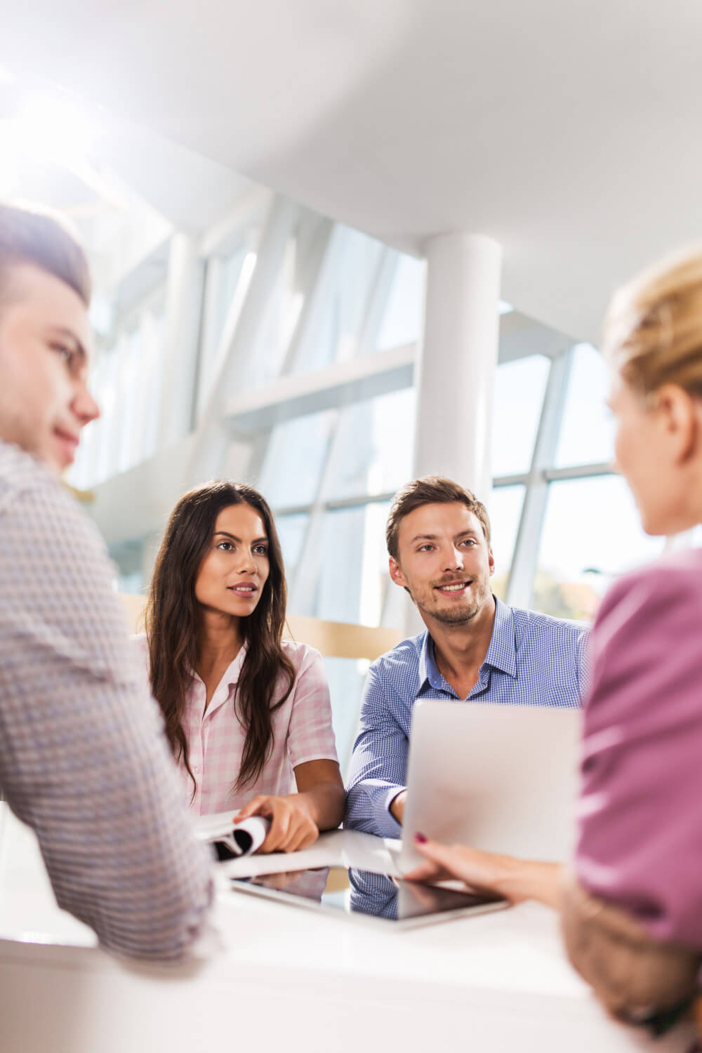 Four coworkers sit around a table, discussing documents and a laptop; two listen attentively while one gestures, in a bright modern office with large windows and white columns.