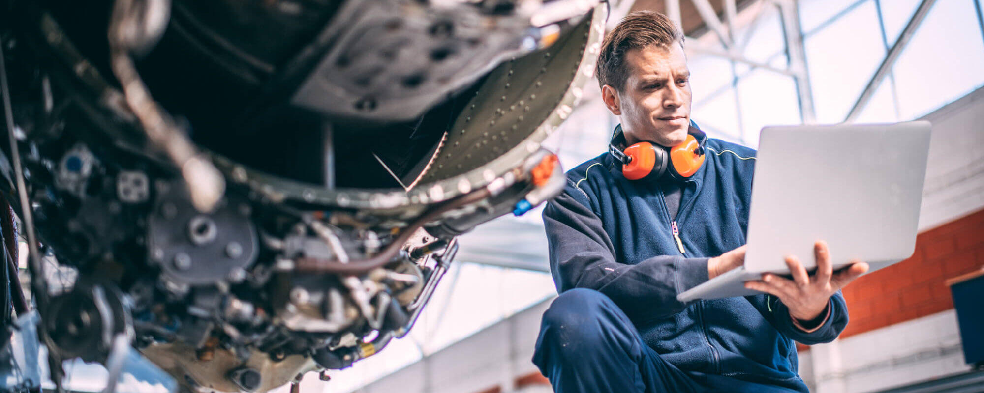 Aircraft technician inspects a laptop while kneeling beside an exposed airplane engine inside a bright hangar, wearing orange hearing protectors and a navy work jacket.