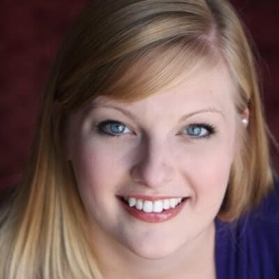 A blonde woman smiles brightly at the camera, revealing teeth; blue-eyed; wearing a purple top; soft lighting and a blurred burgundy background.
