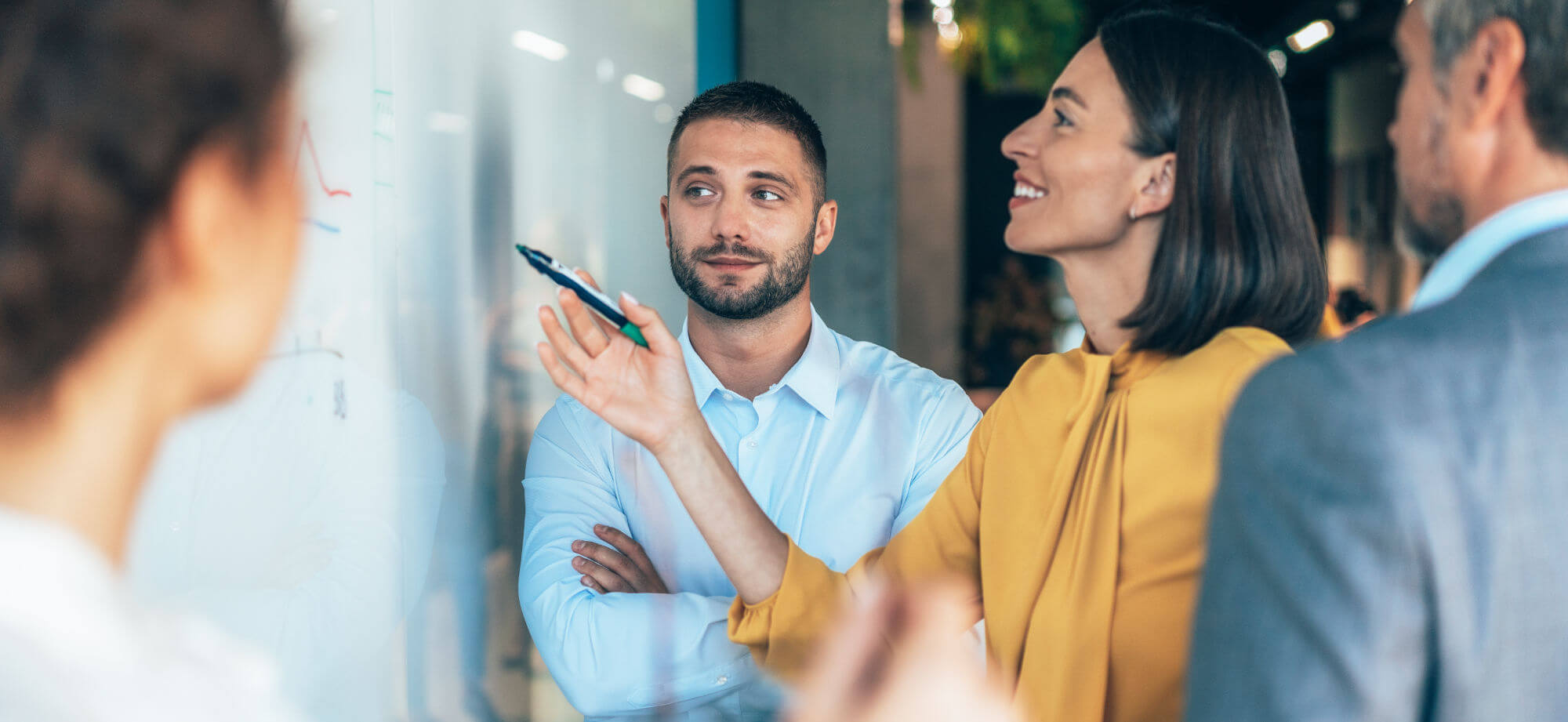 Woman in yellow blouse points with marker at a glass whiteboard; bearded man in light blue shirt watches with arms crossed; two blurred colleagues flank them in a modern office.