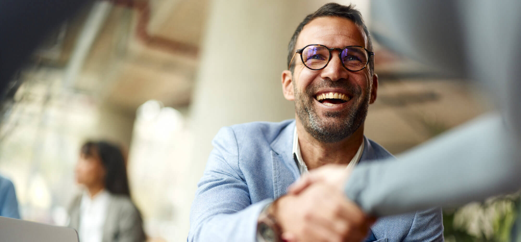 Bearded man with round glasses and light-blue blazer smiles broadly while shaking hands with another person in a bright, blurred office meeting space with colleagues in the background.