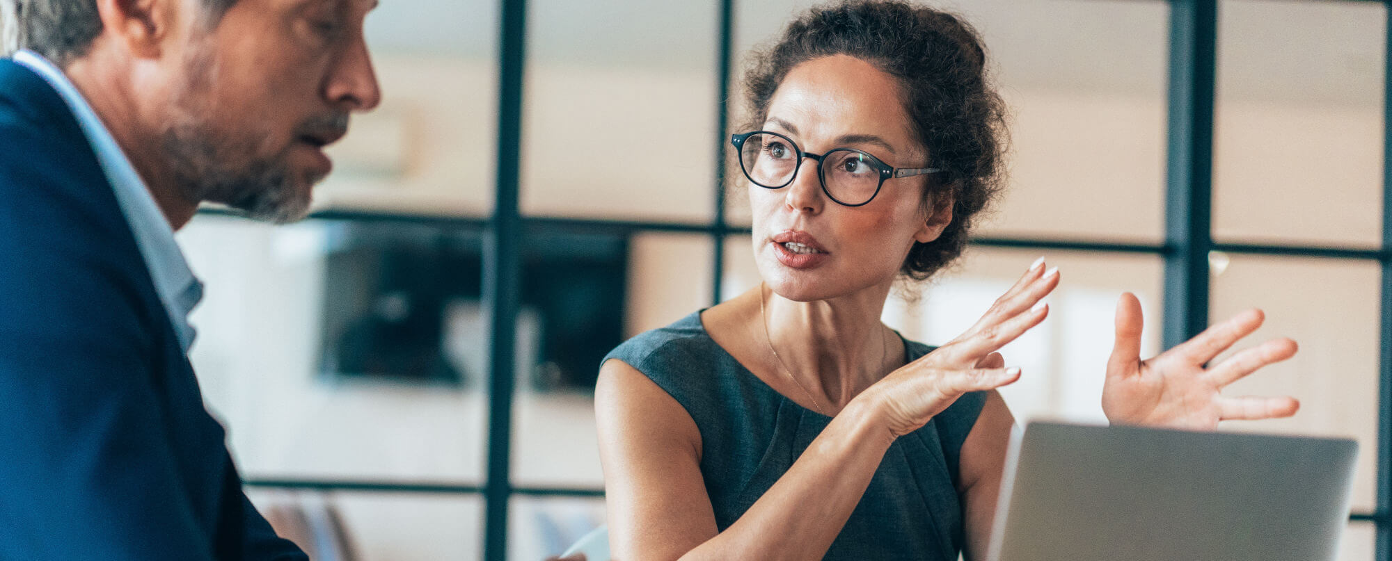 Woman with glasses gestures emphatically while explaining to a male colleague across a laptop, in a modern office with black-framed glass partitions.