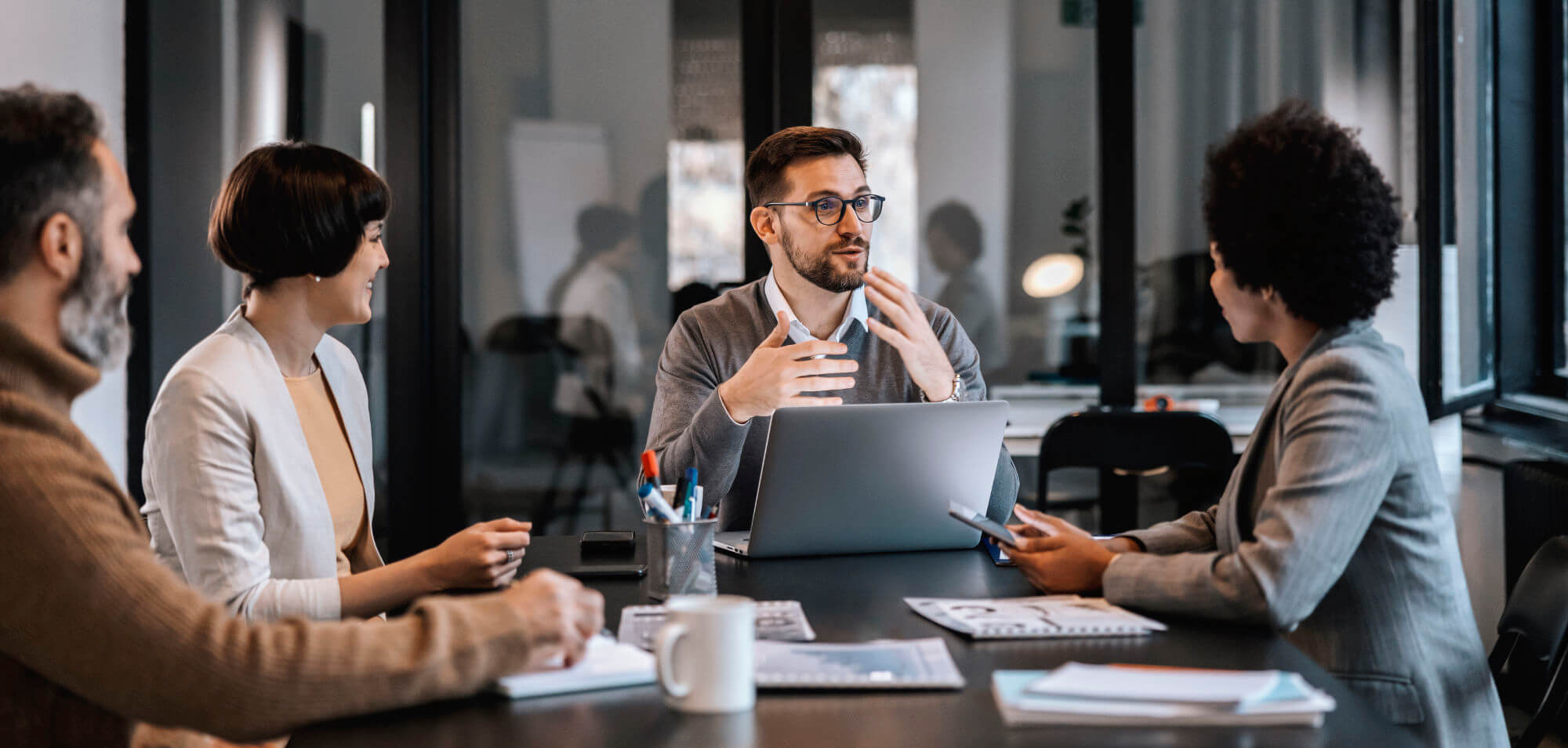 A bearded man wearing glasses gestures while speaking across a laptop, seated at a conference table with three colleagues, notebooks, pens, and a coffee mug in a glass-walled modern office.