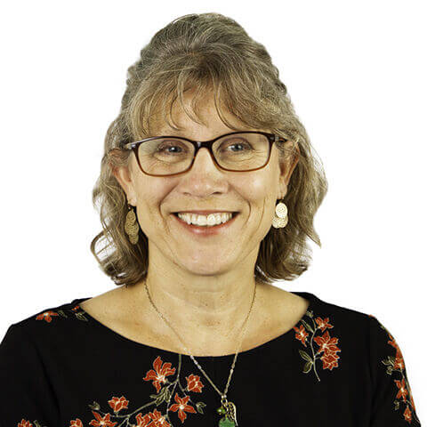 Middle-aged woman wearing glasses and a black floral blouse smiles broadly, showing teeth; short gray hair, dangling earrings and a pendant necklace visible against a plain white background.