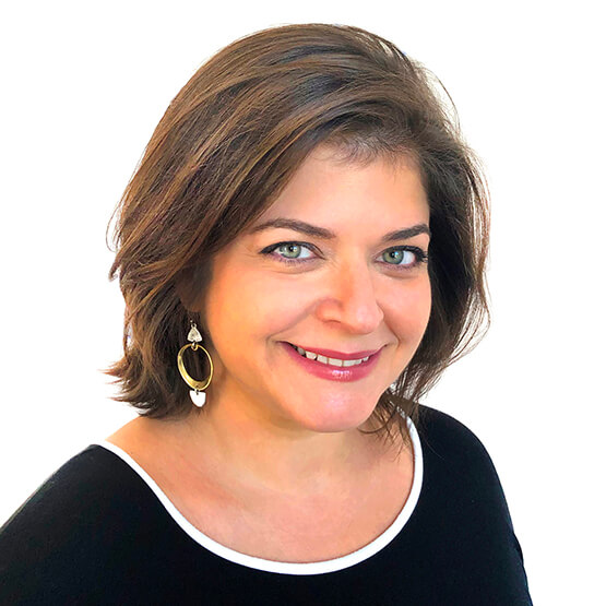 Woman with short brown hair smiles directly at camera, wearing a black top with white trim and large gold hoop-and-heart earrings against a plain white background.