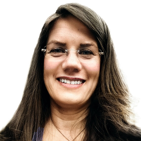 Person with long brown hair and thin-framed glasses smiling directly at the camera in a close-up portrait, showing teeth and a necklace against a plain white background.