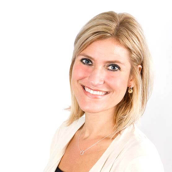Blonde woman smiles directly at camera, wearing a light cardigan, circular pendant necklace and small hoop earrings, posed against a plain white background.