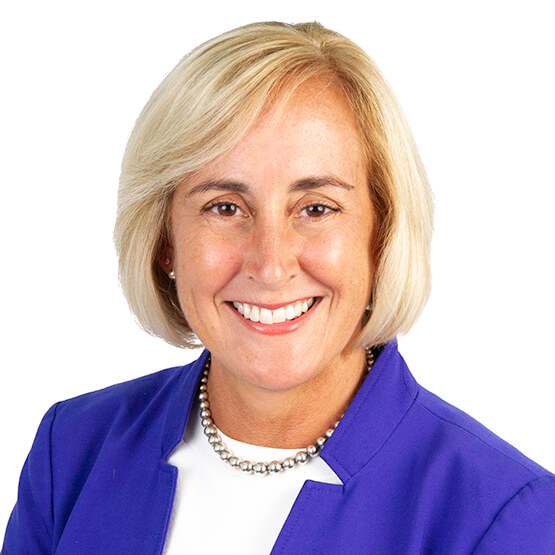 Middle-aged woman smiles at camera in a studio headshot, short blonde hair, wearing a purple blazer, white top and a pearl necklace against a white background.