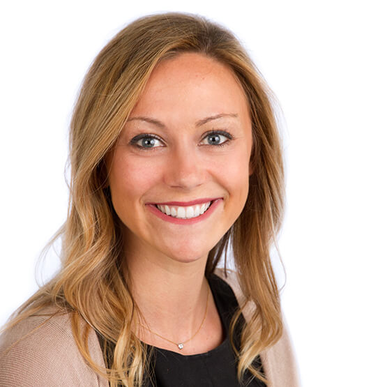 A woman with shoulder-length blonde hair smiling directly at the camera, wearing a black top and light cardigan with a small necklace; close-up studio headshot against a clean white background.