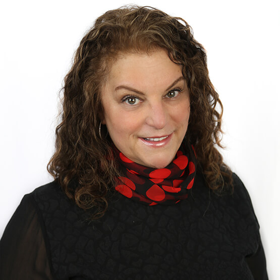 Woman with shoulder-length curly brown hair smiles at the camera, wearing a black top and a black-and-red polka-dot neck scarf, set against a plain white background.