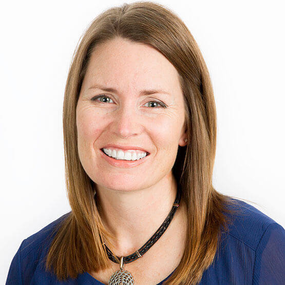 Woman with shoulder-length brown hair smiles at the camera, wearing a blue blouse and a decorative pendant necklace against a plain white background.
