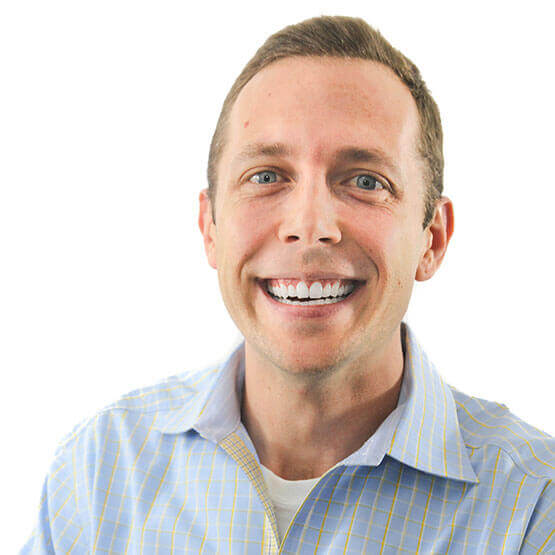 Person smiling broadly, showing teeth; head-and-shoulders pose; wearing light-blue checked shirt over a white tee; plain white background.