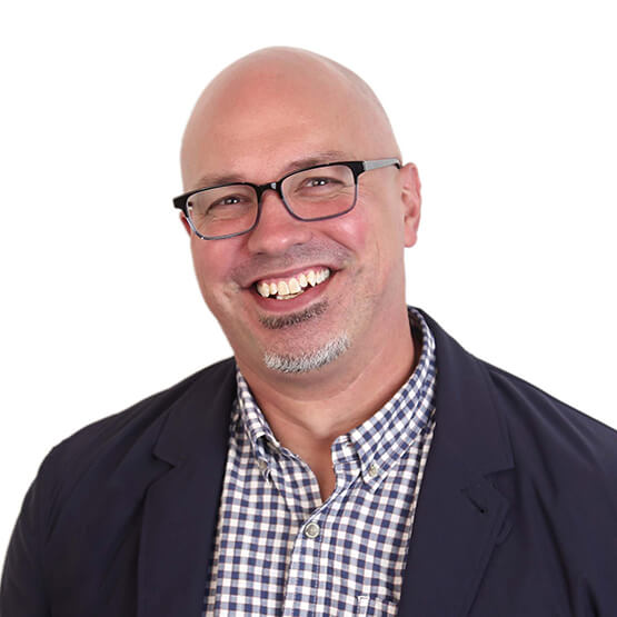 Middle-aged bald man wearing glasses, smiling broadly in a head-and-shoulders portrait against a plain white background; he wears a blue-and-white checked shirt and dark blazer.
