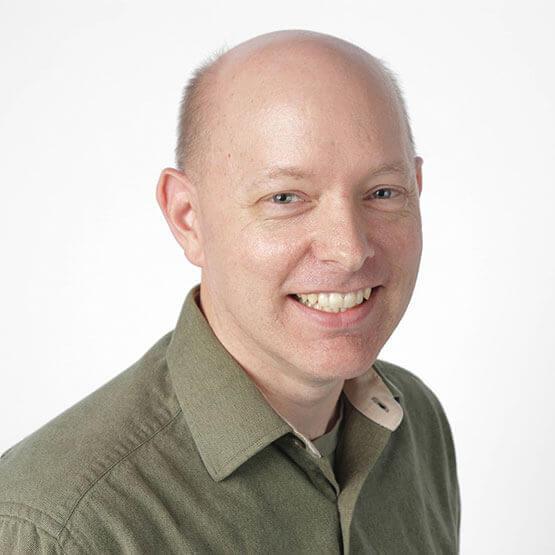A smiling bald man in an olive-green button-up shirt looks slightly to the right; head-and-shoulders portrait against a plain white background.