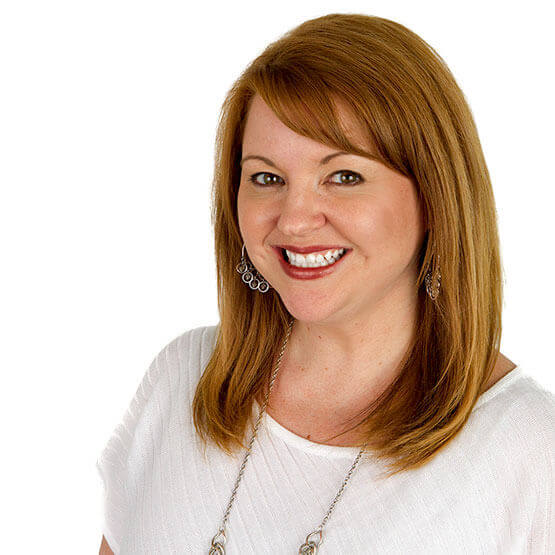 Smiling woman with shoulder-length auburn hair faces the camera, wearing a white top, silver earrings and a long necklace against a plain white background.