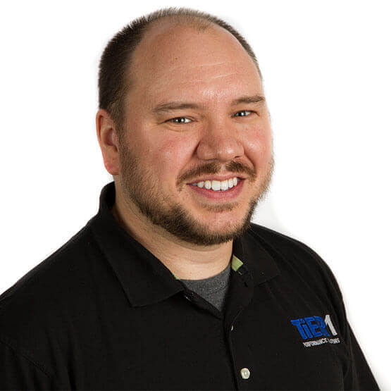 Bearded man smiles at camera, shoulders turned slightly right, wearing a black polo with logo text "TRI Responsive I.T. Services", photographed against a plain white background.