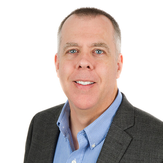 Middle-aged man smiles at camera, wearing a gray blazer over a blue checkered dress shirt, posed shoulders-forward against a plain white background.
