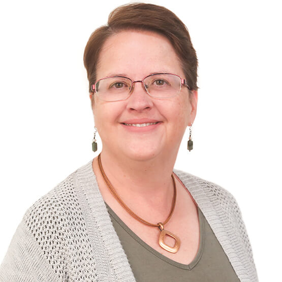 Middle-aged woman smiles directly at the camera, wearing glasses, green top, gray knit cardigan and pendant necklace; short hair and dangling earrings against a plain white background.