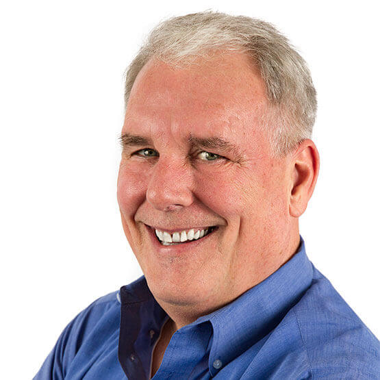 Man with short gray hair smiles broadly, facing slightly left, wearing a blue button-up shirt against a plain white background.