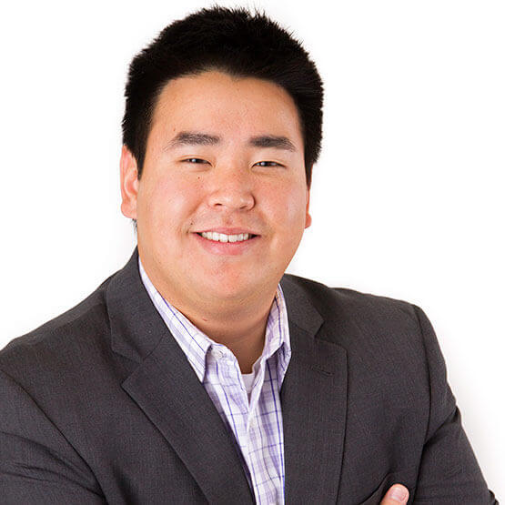 Young man in a gray suit smiles at the camera with arms crossed against a plain white studio background, short black hair and a plaid shirt collar visible.
