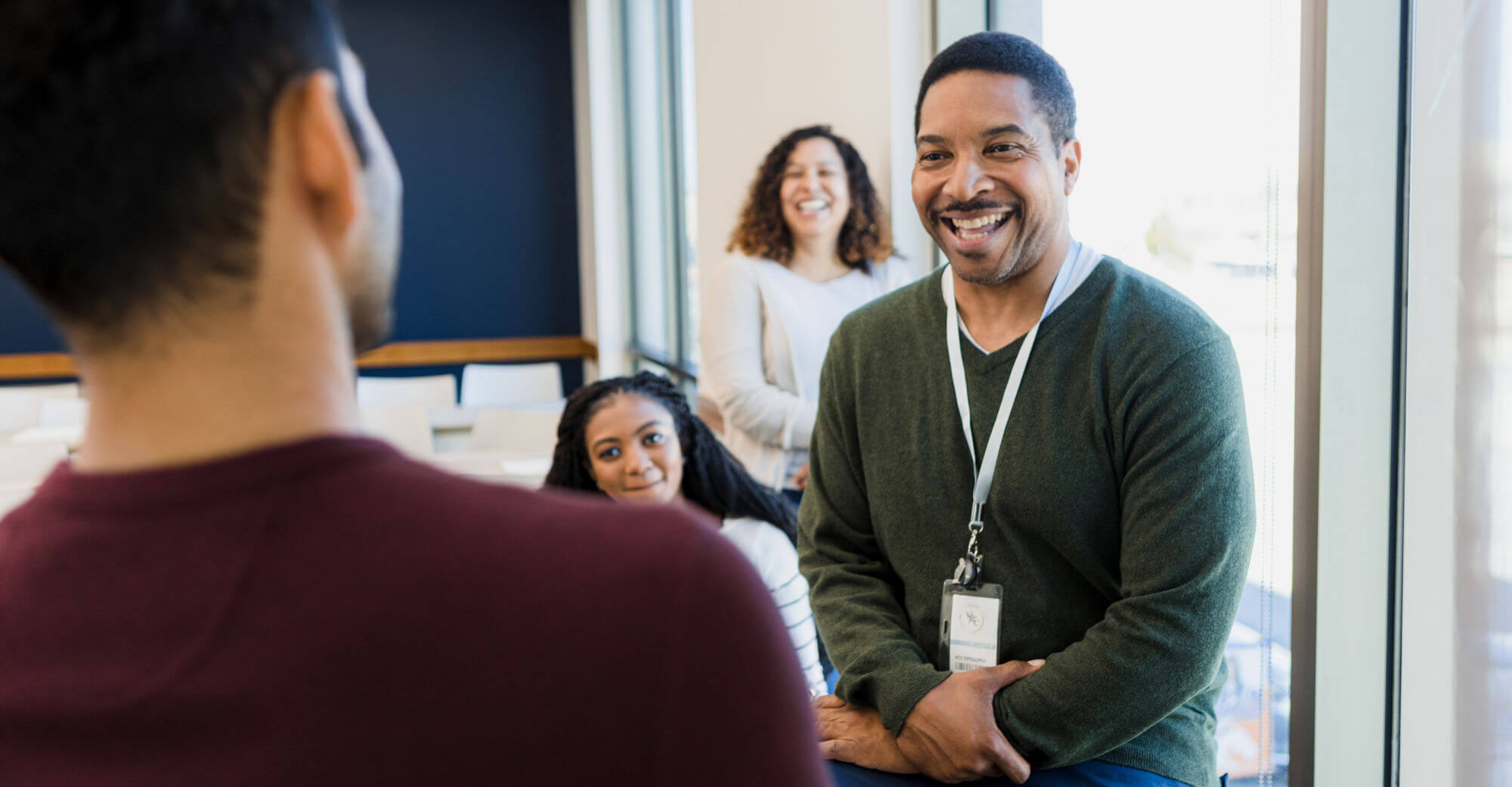 The mature adult male professor smiles and jokes with an unrecognizable young adult male student as the rest of the class enjoys listening in.