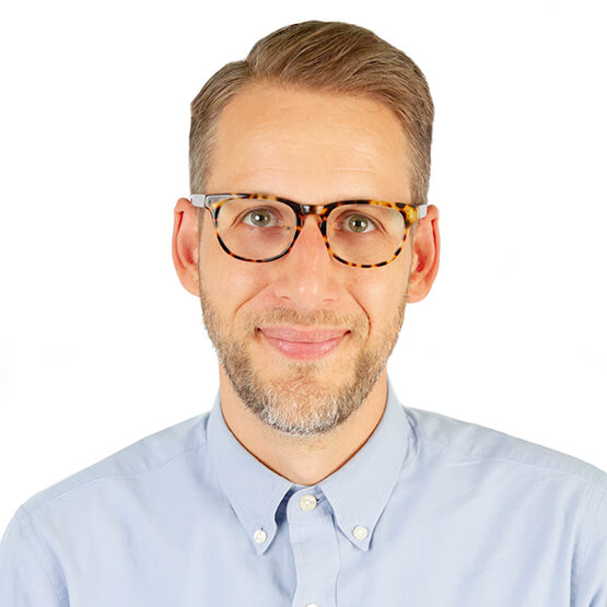 A person with tortoiseshell glasses and a light blue shirt, smiling subtly.