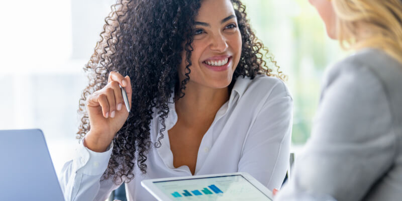 Two businesswomen smiling and discussing data on a tablet.