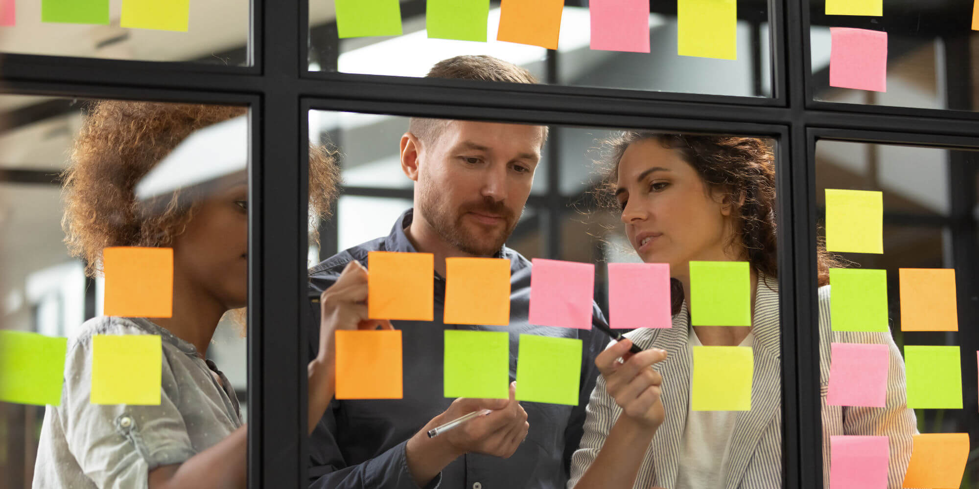 3 businesspeople working together while writing on sticky notes and putting them on a glass wall.