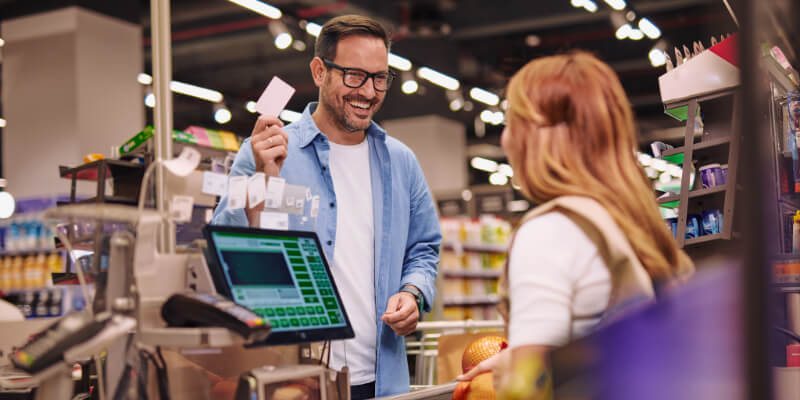 A cheerful man customer interacts with a woman cashier at a supermarket checkout. Fresh produce items are on the conveyor, showcasing a grocery shopping experience.