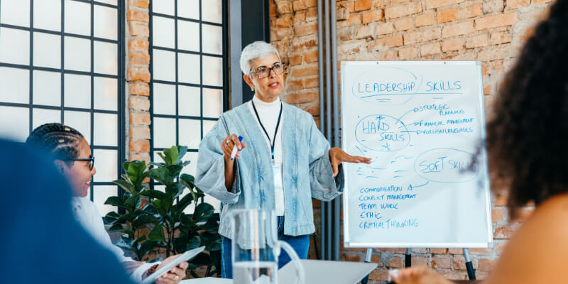 A mature businesswoman is presenting a strategy on leadership skills to engaged, diverse colleagues in a fresh and innovative office setting.