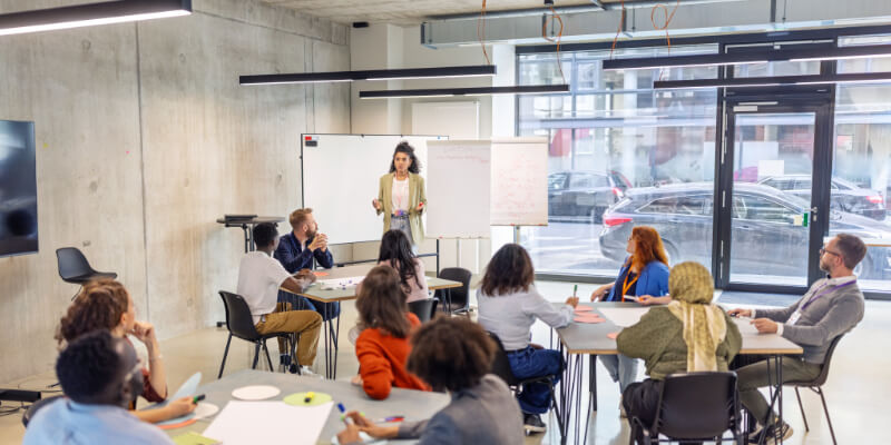 Female professional standing by a flipchart, addressing businesspeople seated in small groups at different tables. Highlights her leadership skills and interactive approach during a seminar at a convention center.