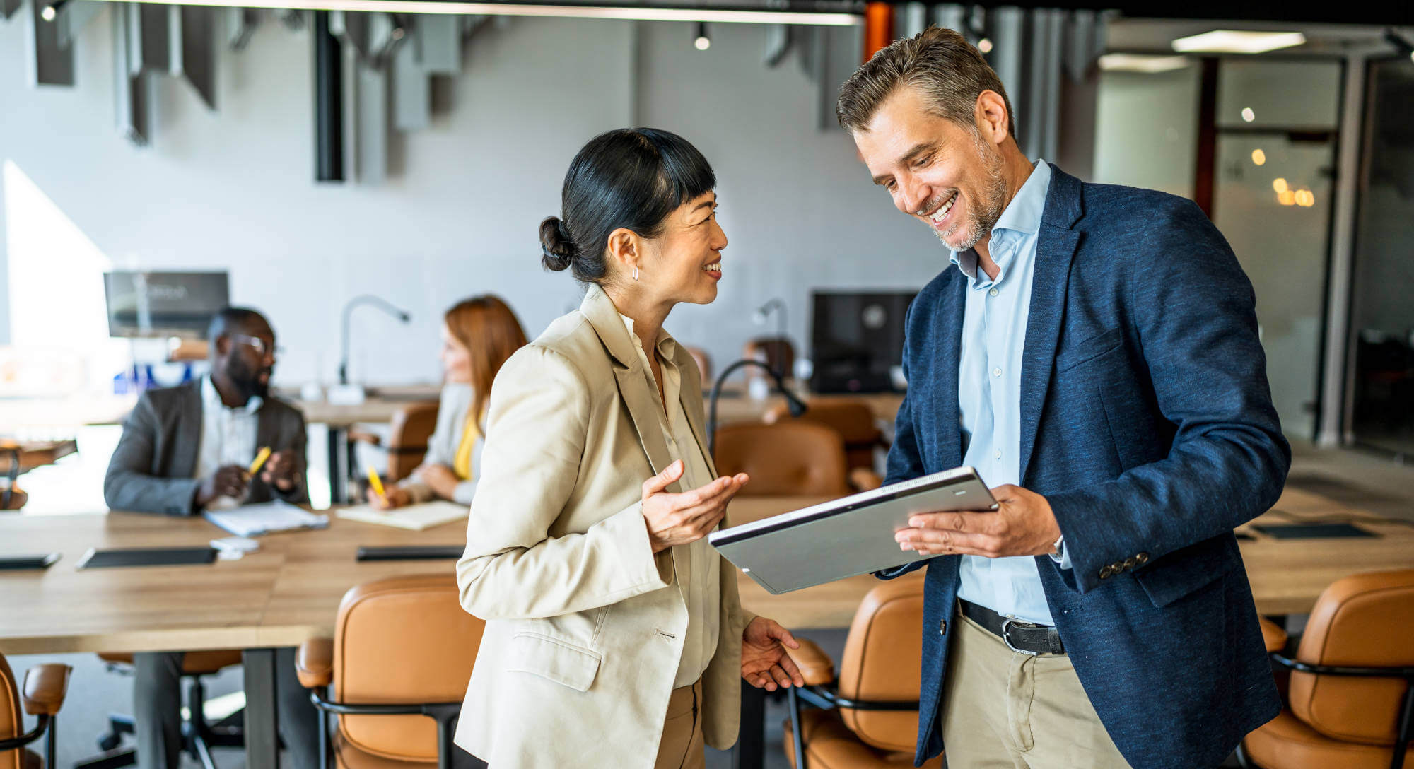 Two colleagues smiling and discussing something on a tablet in an open office.