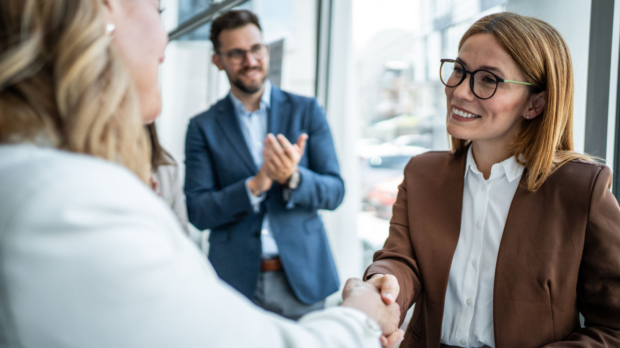 Two colleagues shaking hands while another person applauds in the background.